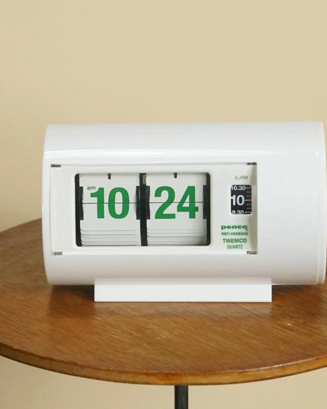 A white flip-flap digital clock with green numbers displaying 10:24, sitting on a round wooden table against a beige wall.