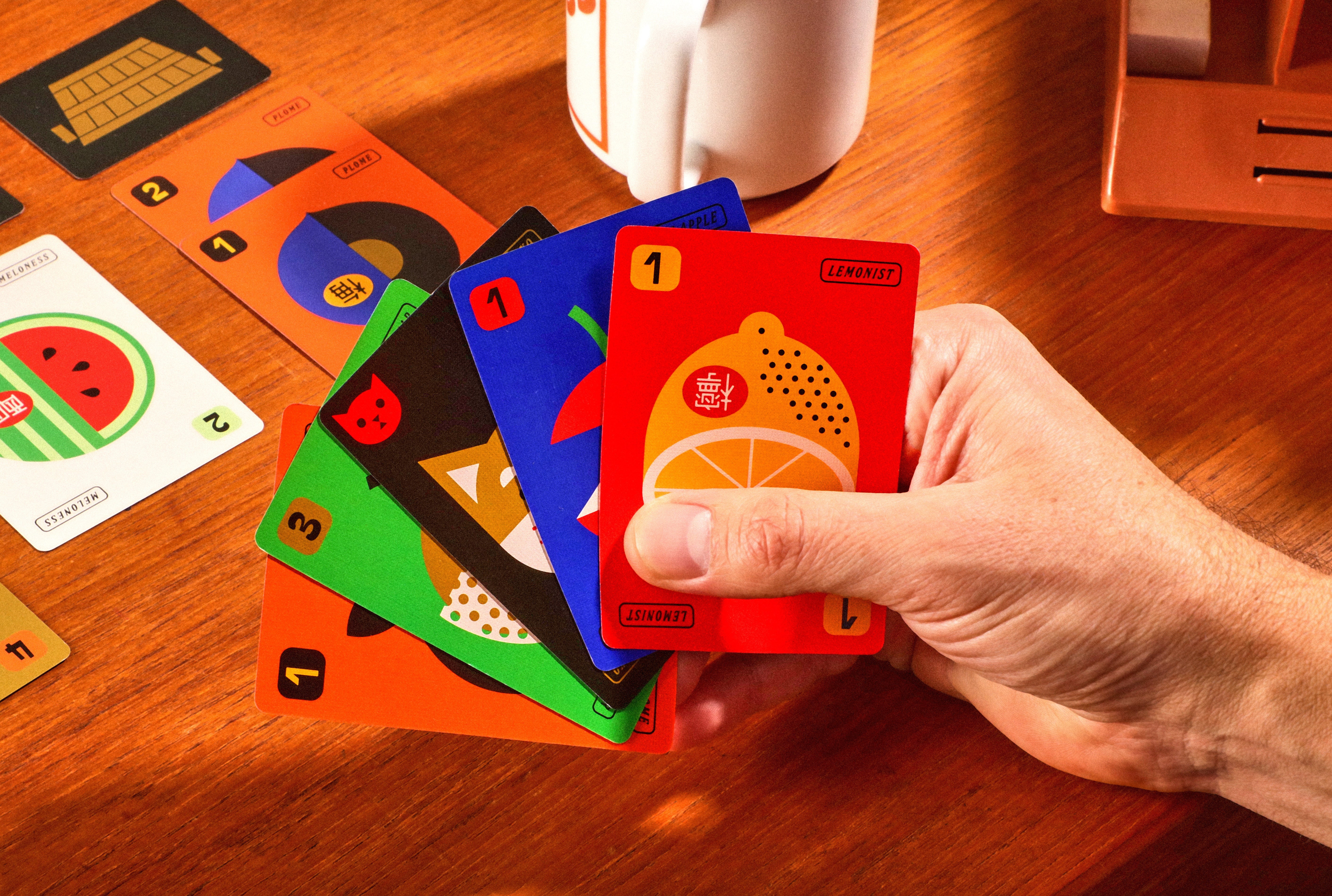 Hand holding colorful playing cards on a wooden table with a white mug in the background.