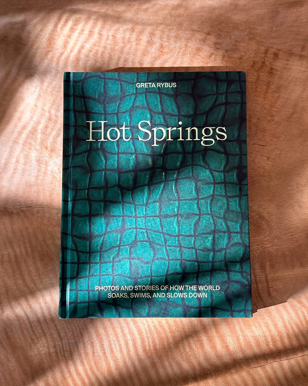 The book 'Hot Springs' by Greta Rybus lies flat on a textured, warm brown surface with soft diagonal shadows. The cover features a dark teal and black reptile-like pattern with white text.