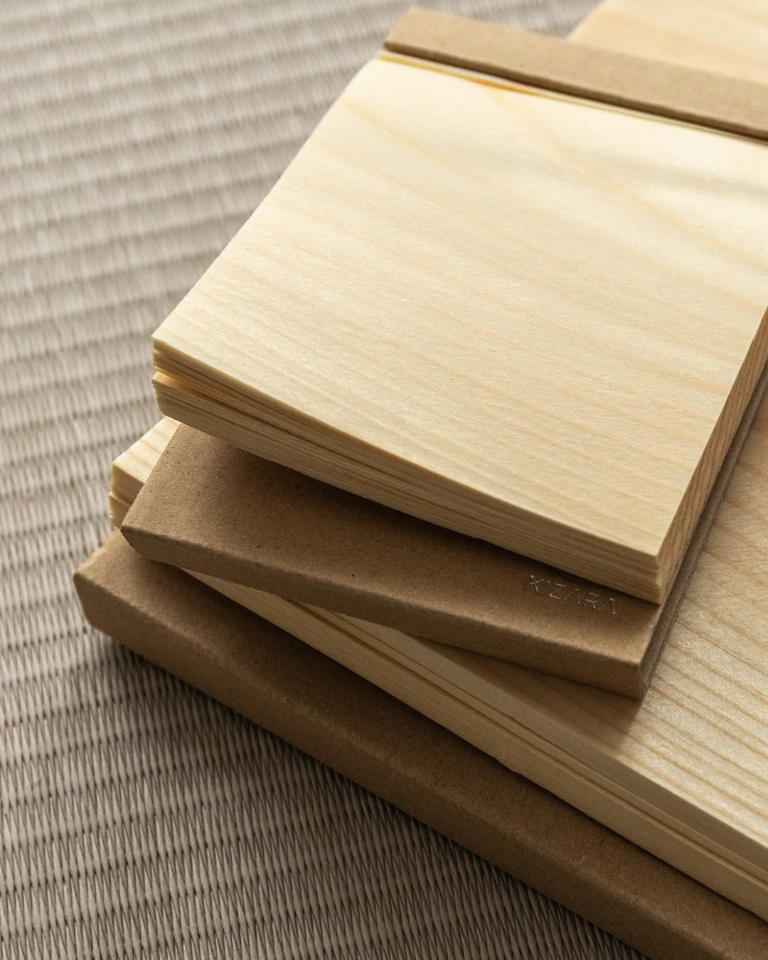 Stacked sheets of light natural wood and brown kraft paper on a woven beige fabric background.