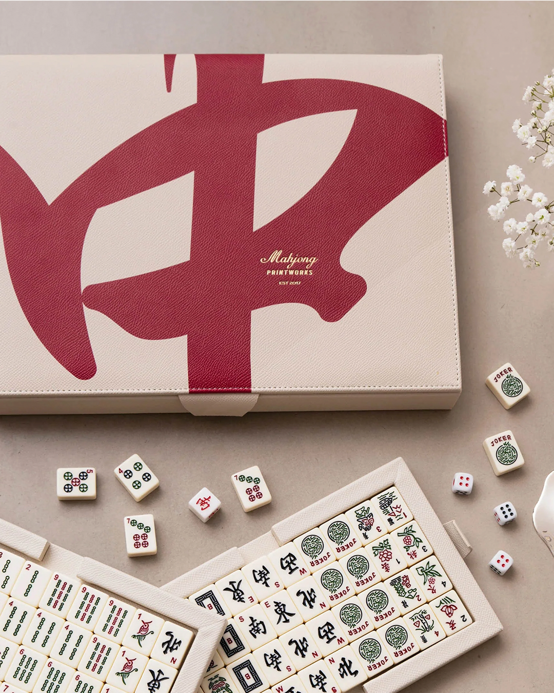 A Mahjong set with a cream-colored carrying case featuring a large red Chinese character, shown alongside several open tiles and dice on a neutral beige surface.