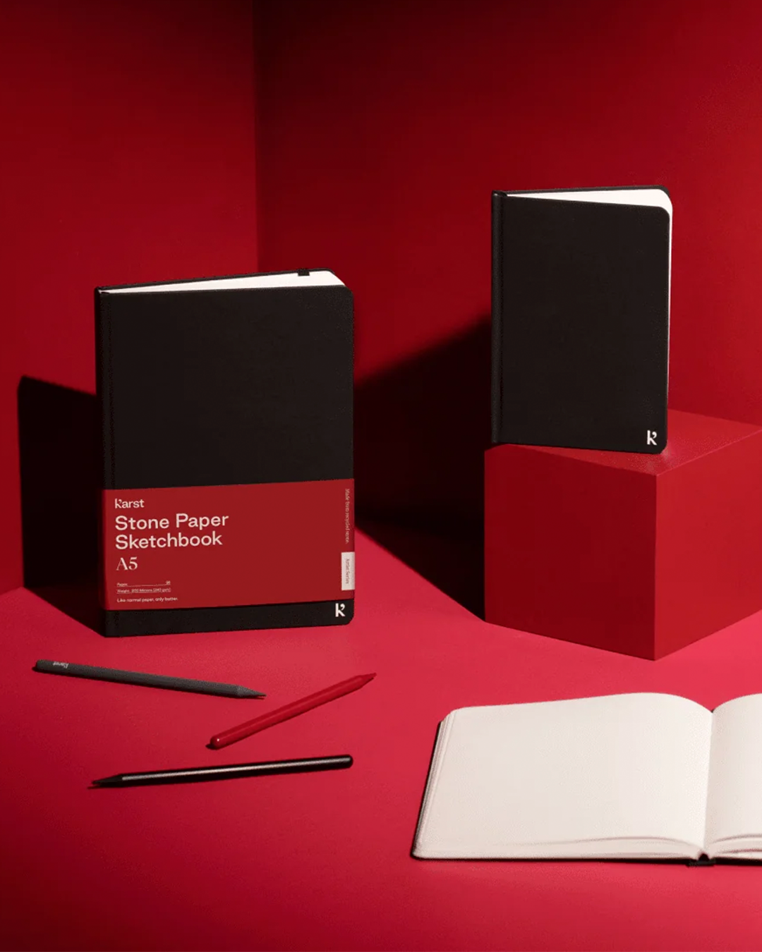 A Barst Stone Paper Sketchbook AS in a matte black cover with a red front label, shown alongside an open blank sketchbook, two pens, and a red display block against a red studio background.