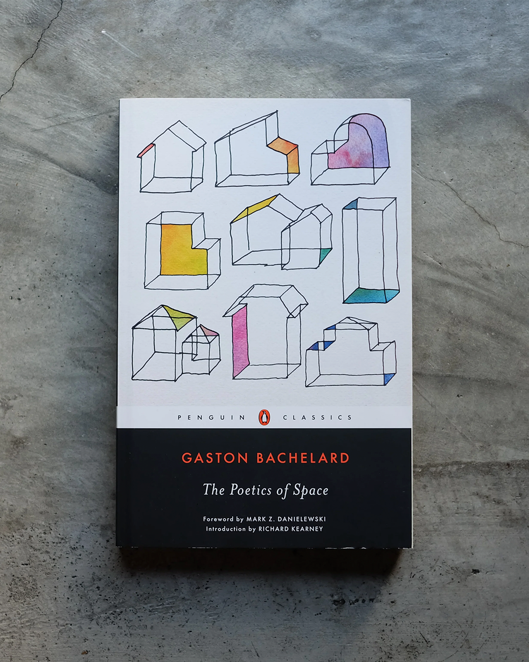 The book cover of Gaston Bachelard's 'The Poetics of Space' by Penguin Classics, featuring minimalist line drawings of geometric house forms on a white upper section and a black lower section, photographed flat on a textured gray concrete surface.