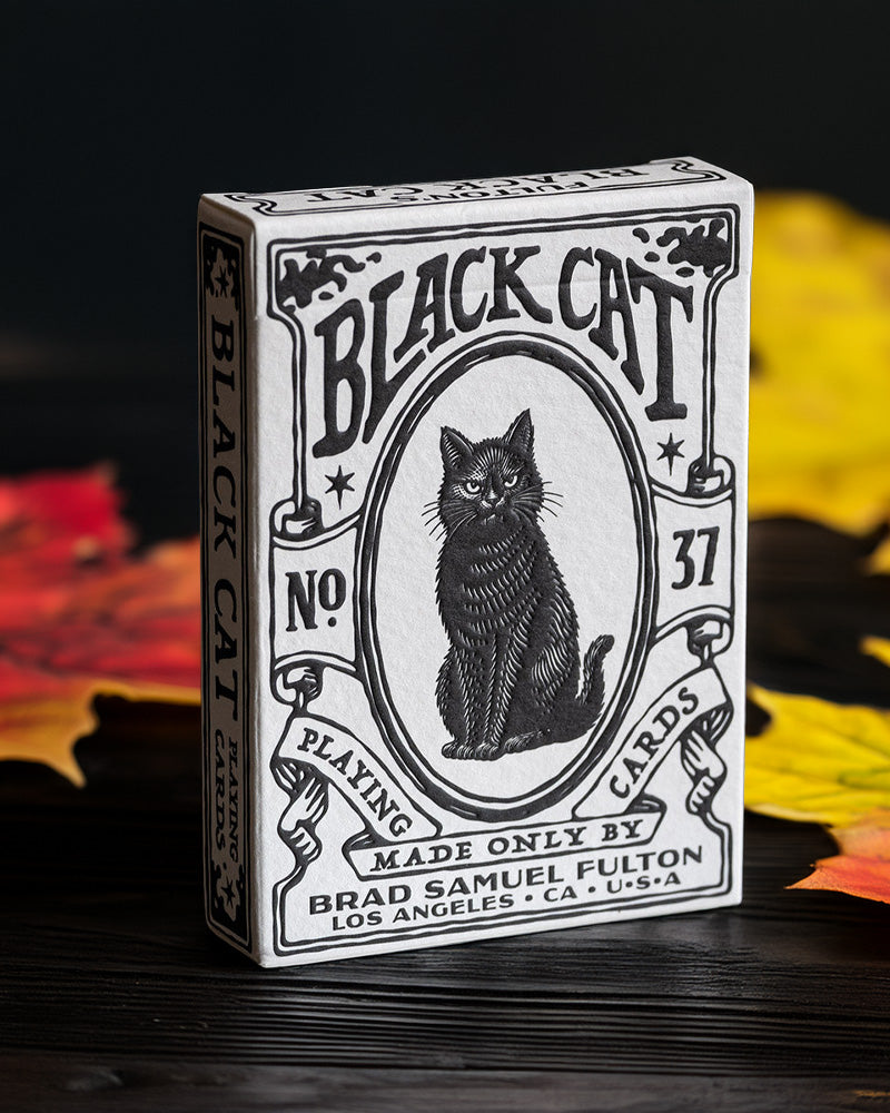 A boxed deck of 'Black Cat' playing cards stands upright on a dark wooden surface with autumn leaves blurred in the background. The box is white with black vintage-style illustration and text, featuring a cat portrait in the center.