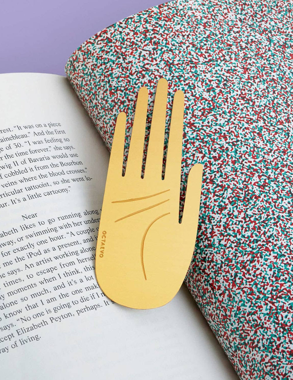 A gold hand-shaped utensil with four prongs, embossed with the word 'grace', resting on an open book next to a roll of multicolored glitter paper on a light purple surface.
