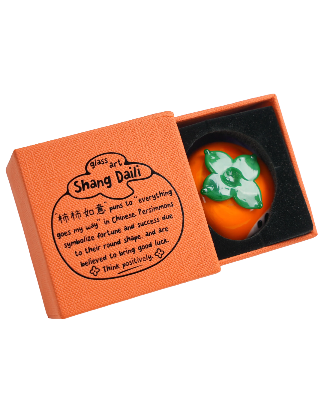 An orange textured gift box shown open, with a black interior insert holding a round persimmon figurine featuring a green floral design. The lid has printed Chinese characters and the words 'glass art Shang Dalli'.