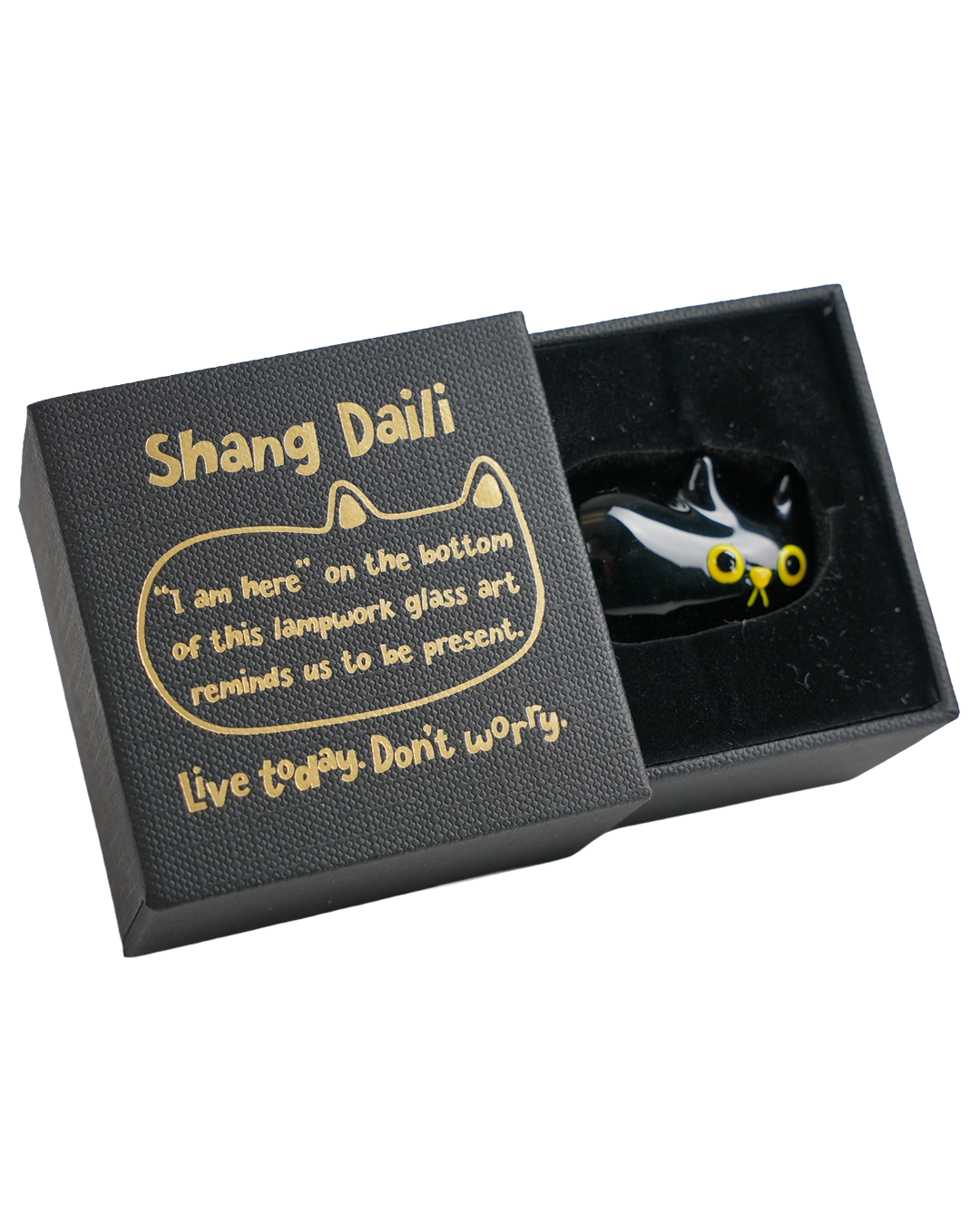 A black cat-shaped glass lampwork art piece with yellow eyes displayed inside an open black gift box. The box lid features gold text reading 'Shang Daili' and a quote about lampwork glass art.