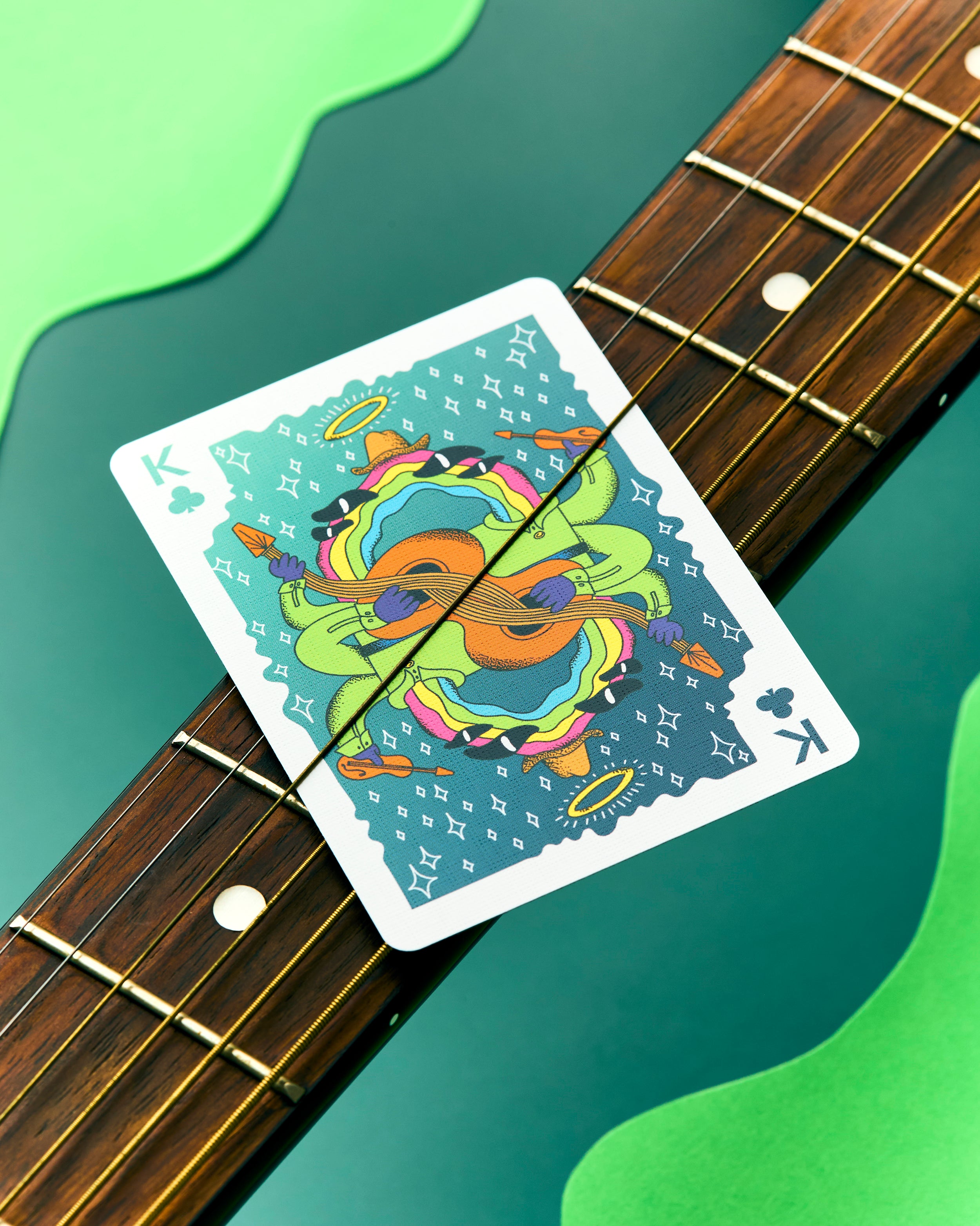 A playing card with a colorful psychedelic guitar illustration lies diagonally across the neck of an electric guitar against a green background.