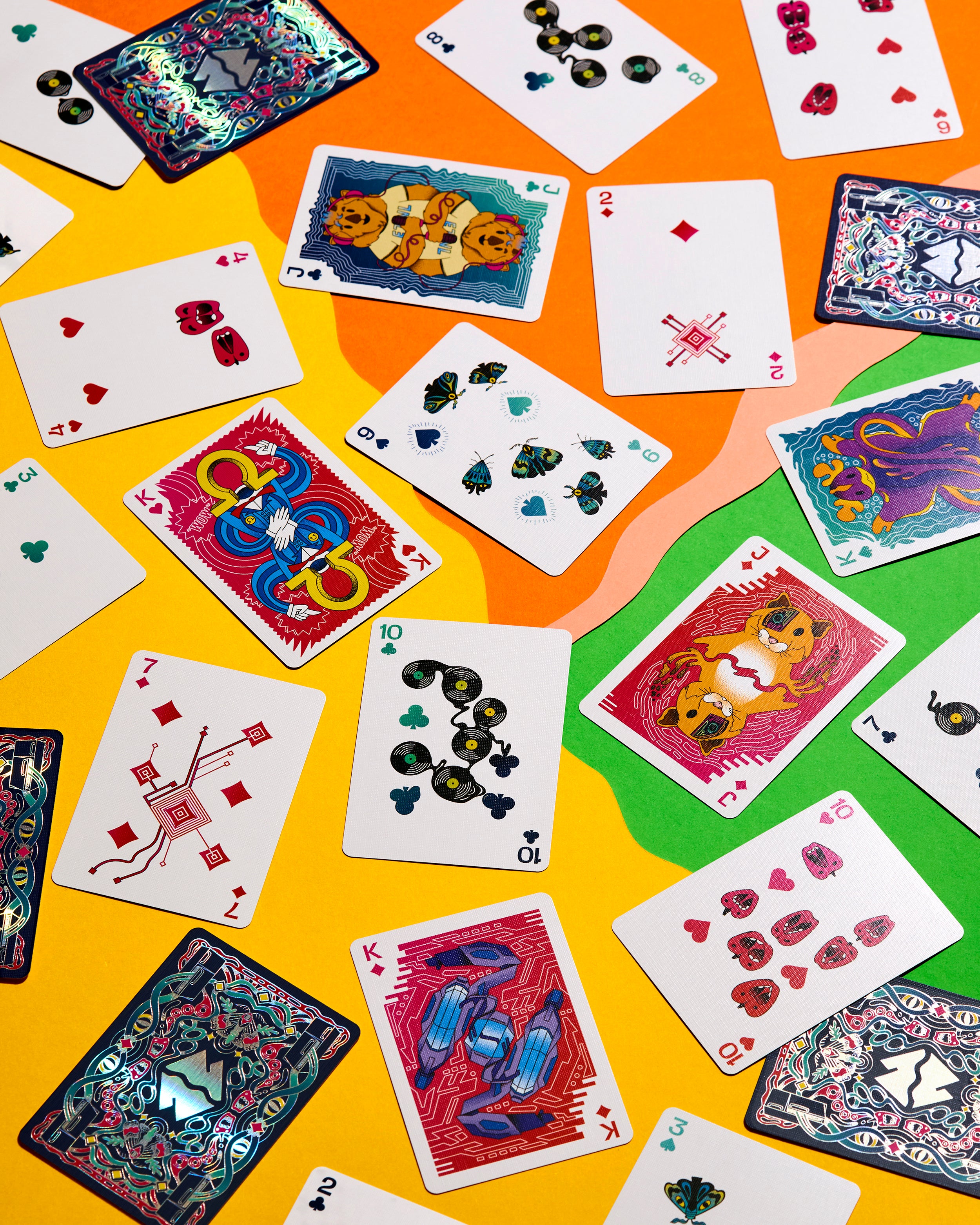 A flat-lay arrangement of various playing cards spread across a vibrant, abstract background of orange, yellow, and green shapes. The cards feature colorful, artistic illustrations of animals, hearts, suits, and decorative patterns on their faces, and show detailed, multicolored back designs.