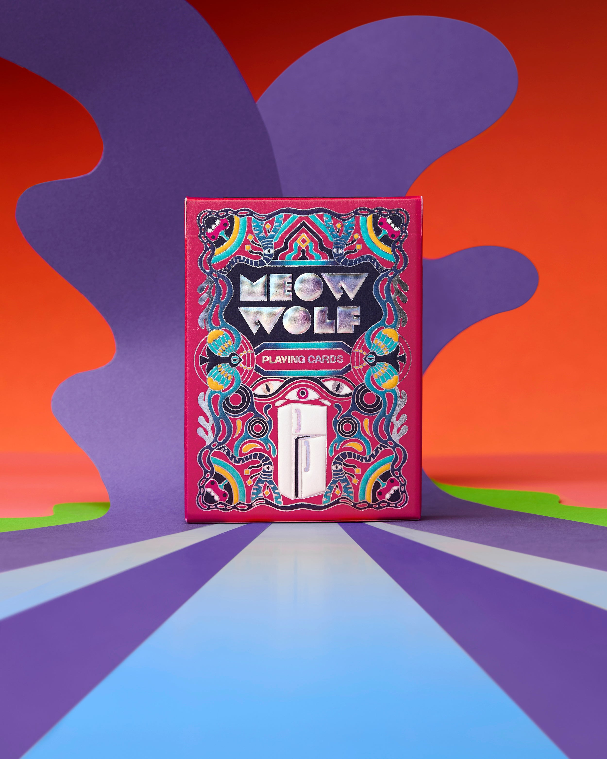 The 'Meow Wolf' Playing Cards deck box stands upright on a colorful, abstract background. The box features a vibrant, psychedelic design with rainbow patterns and the brand name 'MEOW WOLF' in bold white letters.