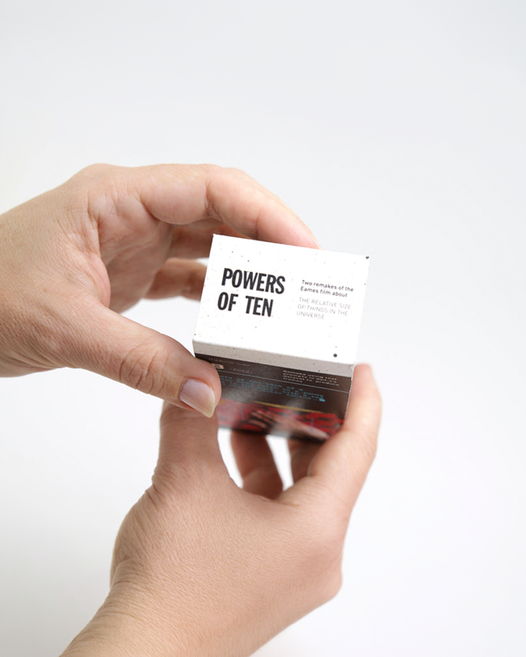 A person's hands holding a small white book or card titled 'POWERS OF TEN' against a clean white background.