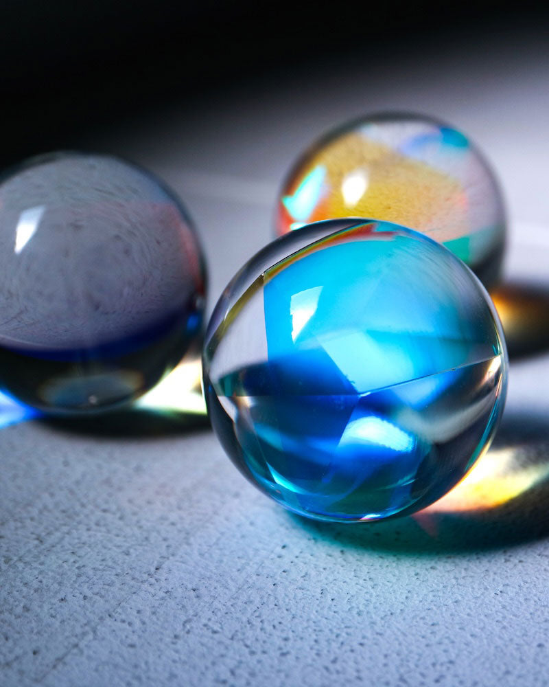 Three transparent glass or acrylic decorative spheres with iridescent rainbow reflections, photographed on a textured light surface with dramatic lighting.