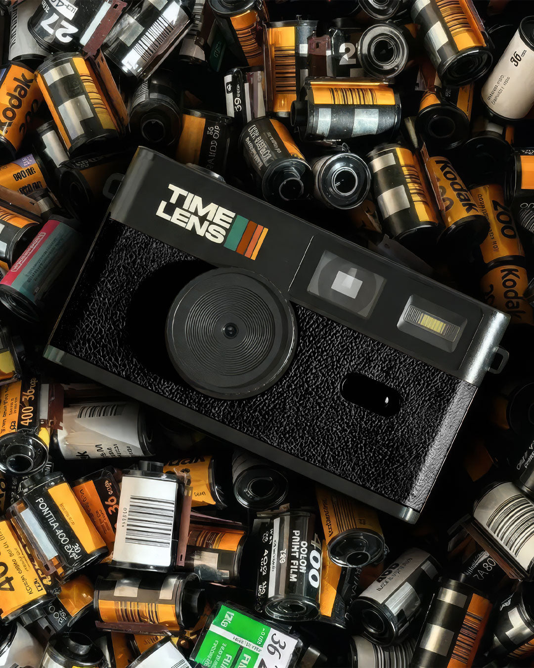 A black Time Lens instant camera lies on top of a dense pile of vintage film canisters and rolls, many showing Kodak branding.