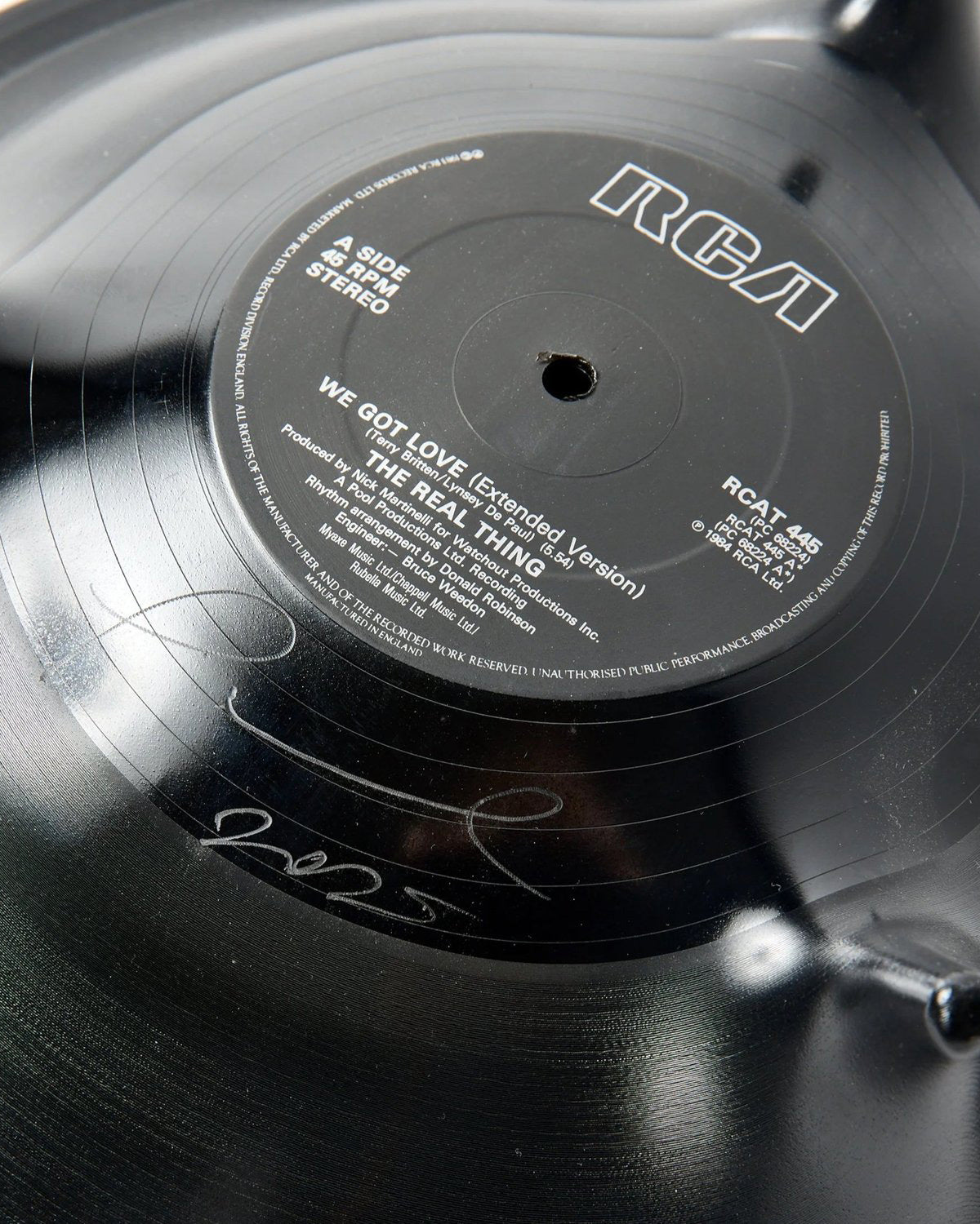 Close-up of a black vinyl record with the RCA logo and white text reading 'We Got Love (Extended Version)' and 'The Real Thing,' showing part of a signature on the lower left side.