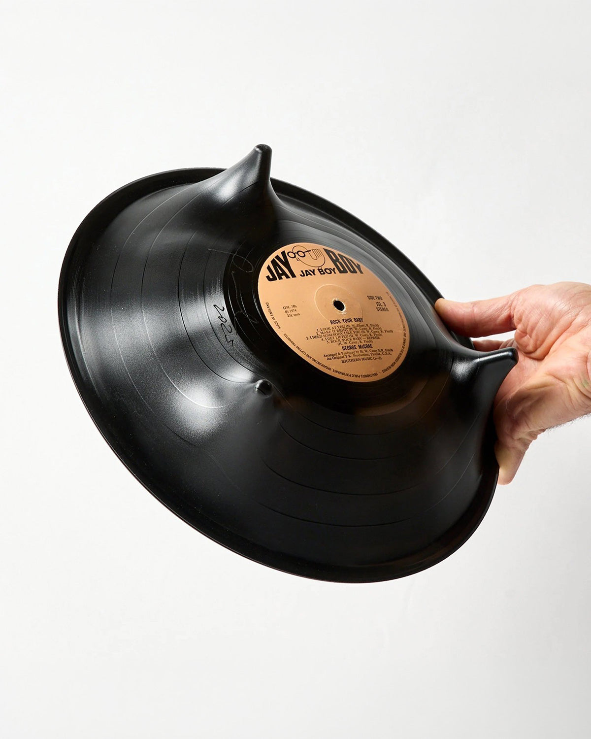 A hand holding a black vinyl record shaped like a bowl, featuring a tan center label with 'JAY BOY' text, against a plain white background.