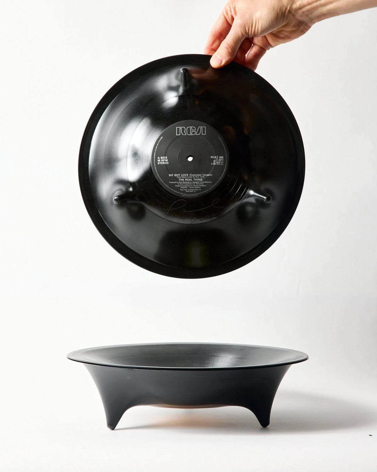 A black molded vinyl record bowl with a three-legged base, shown with a hand holding the bowl from above while displaying its underside against a white studio background.