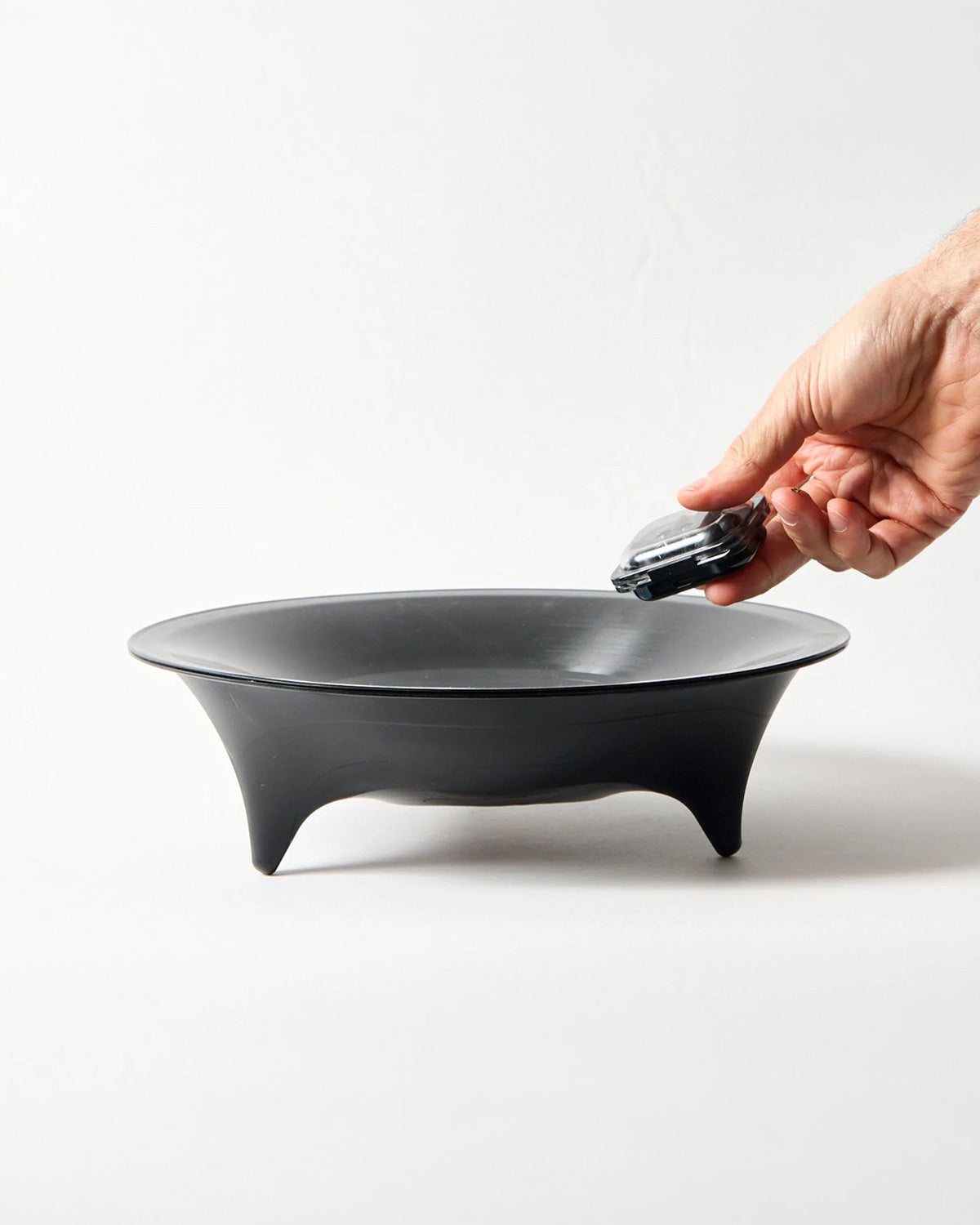A black bowl with three legs , shown on a white studio background with a hand holding a small black object above it.