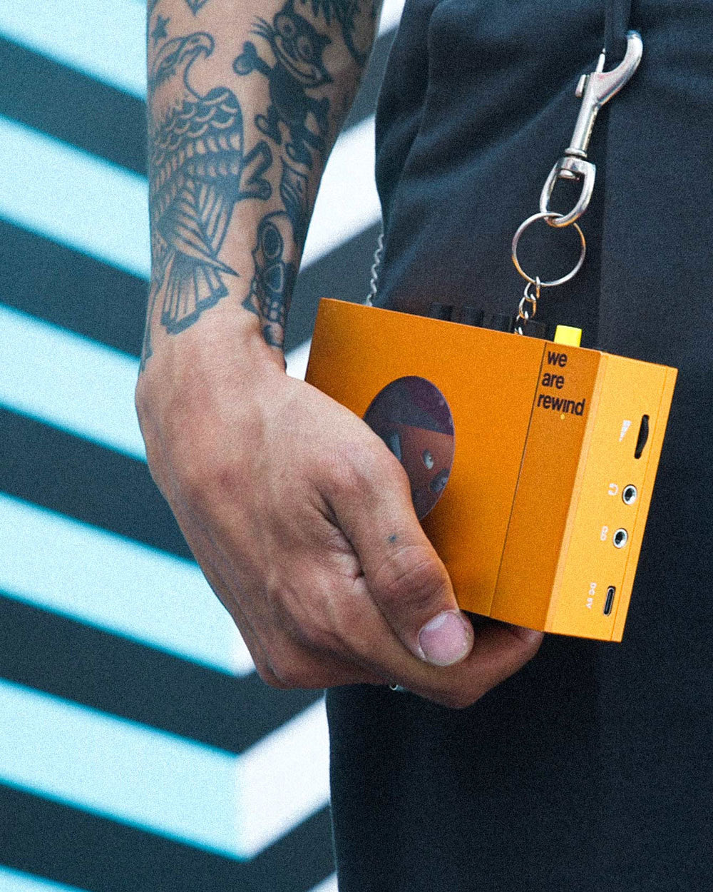 A tattooed person holding a retro cassette player hanging from their belt loop