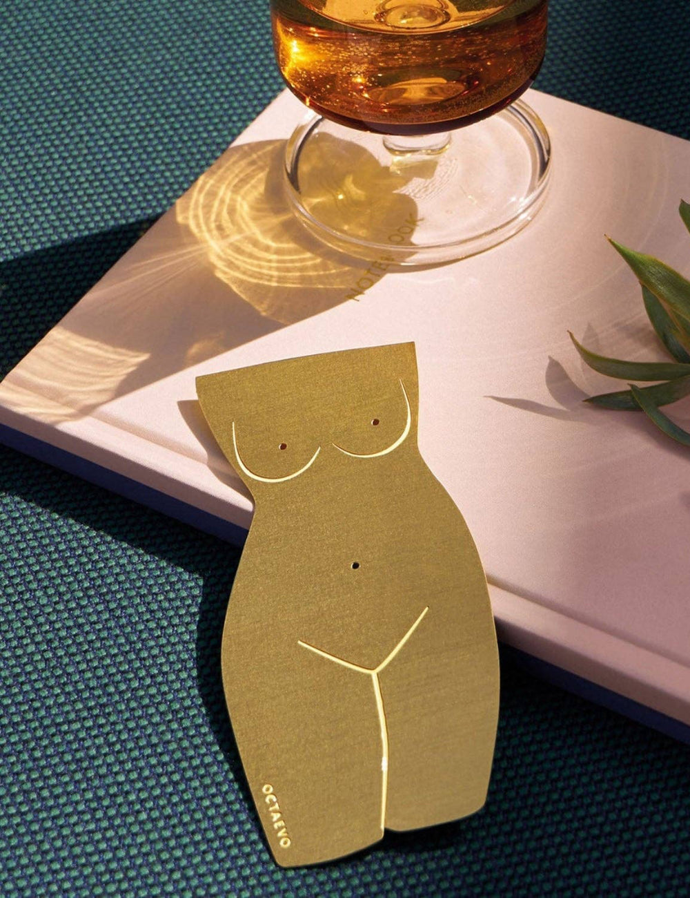 Gold female torso-shaped object on a surface with a glass of amber liquid and plant in the background.