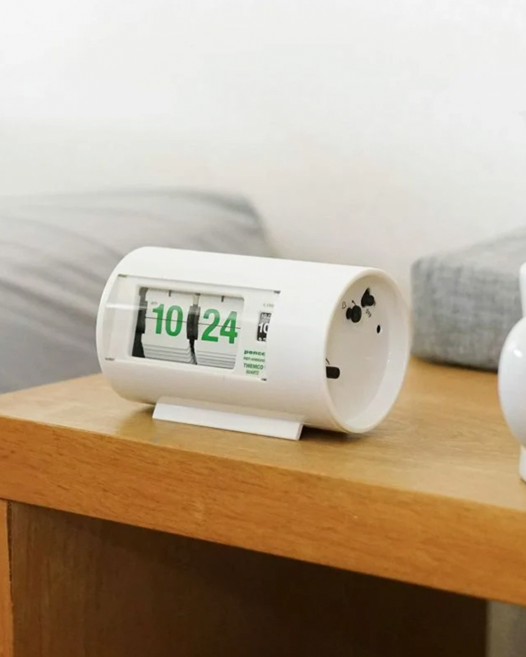 A white cylindrical digital alarm clock with green numbers, resting on a light wood bedside table in a softly lit room.