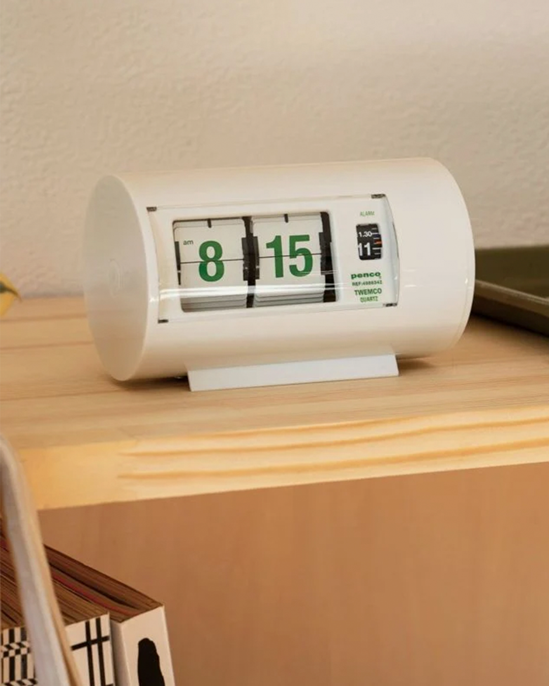 A white cylindrical flip clock with green numbers sits on a light wood shelf in a home setting.