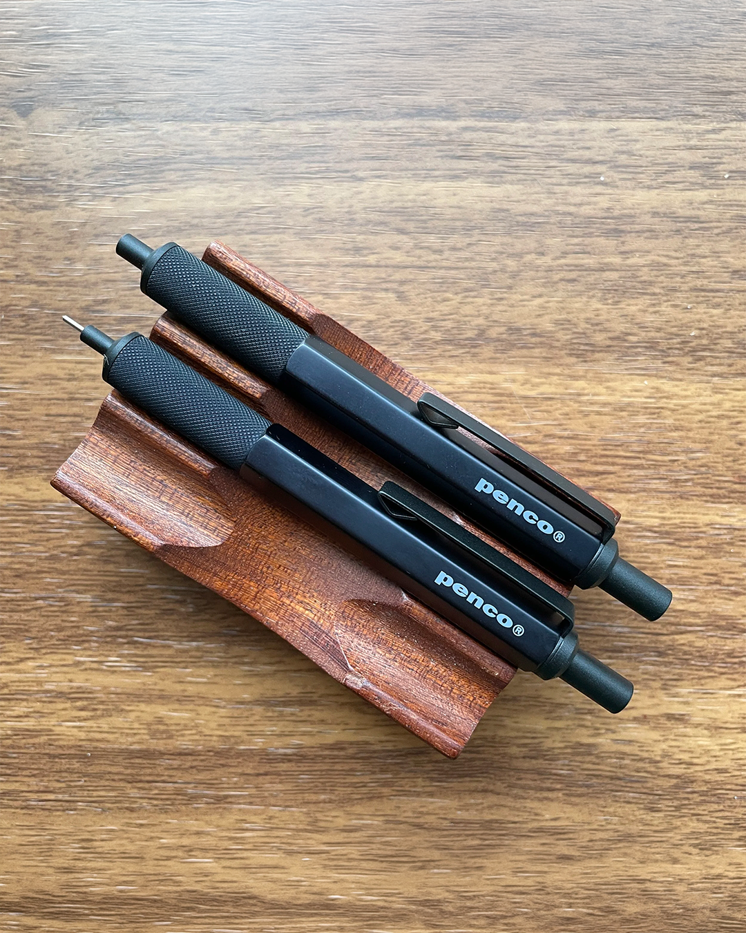 Two black Benco mechanical pencils with textured black grips rest on a carved wooden stand, set against a warm brown wooden tabletop background.