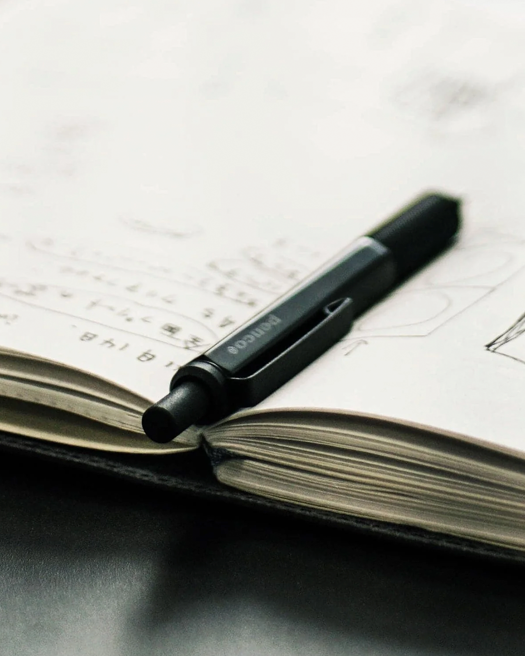 A black Pilot ballpoint pen resting on an open notebook with handwritten and sketched pages.