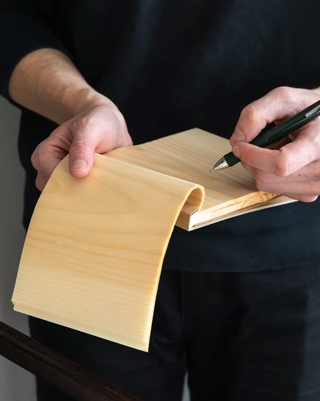 A close-up of a person's hands holding a flexible, light-colored wooden panel or book while writing on the cover with a black pen. The person is wearing a black shirt, and the background is a dark, out-of-focus interior.