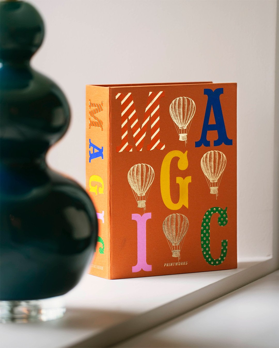 A square orange book or box standing on a white windowsill, decorated with large multicolored letters and hot air balloon illustrations. A dark glass vase is blurred in the foreground on the left.