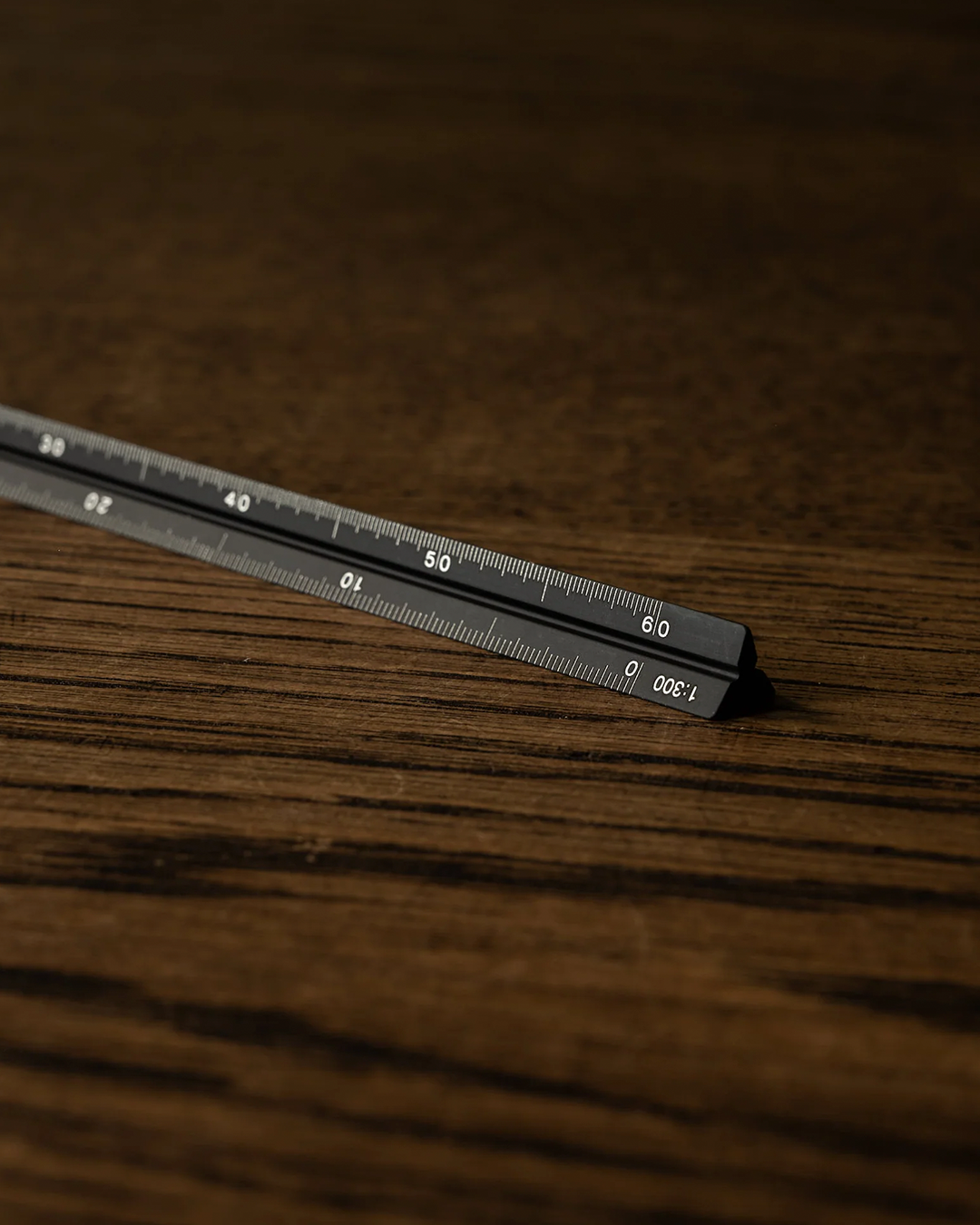 A black metal ruler with white printed measurement markings, shown diagonally on a dark brown wood grain surface.