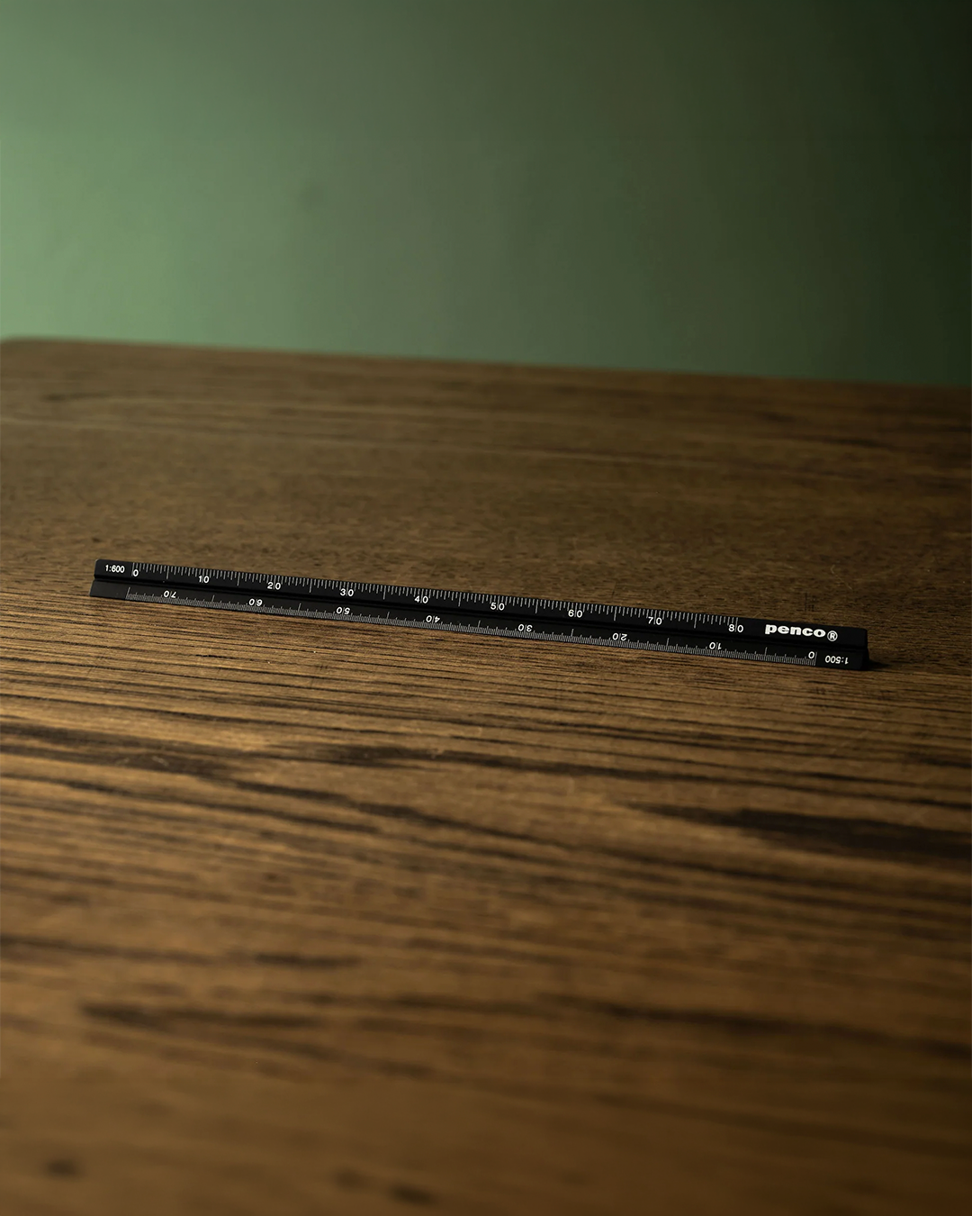 A black plastic ruler with white markings and the 'pencos' brand name, resting on a dark wood tabletop against a soft green background.