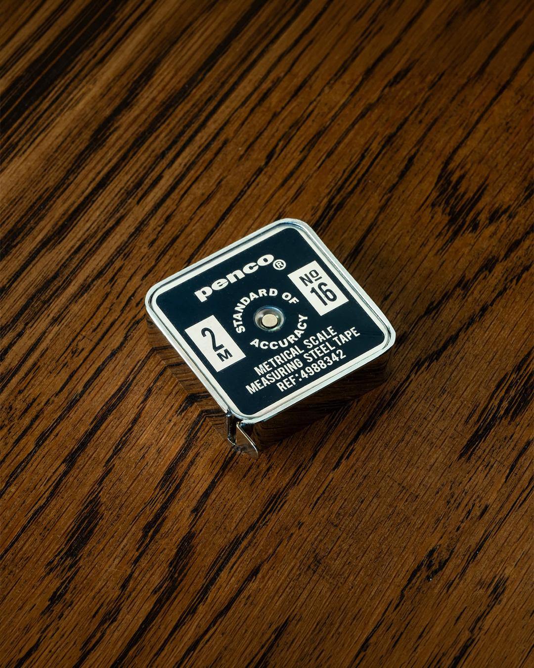 A Penco 2-meter steel tape measure with a silver metal casing and a dark blue label, resting on a dark wood grain surface.
