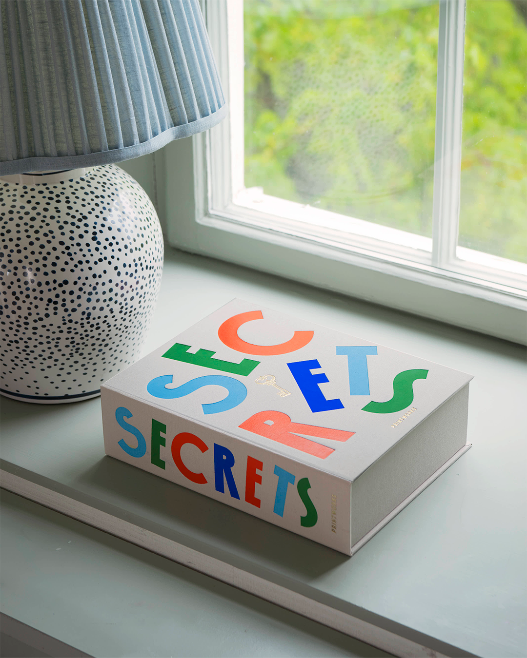 A white hardcover book titled 'SECRETS' with colorful, playful lettering sits on a windowsill next to a patterned lamp and a window looking out to green foliage.