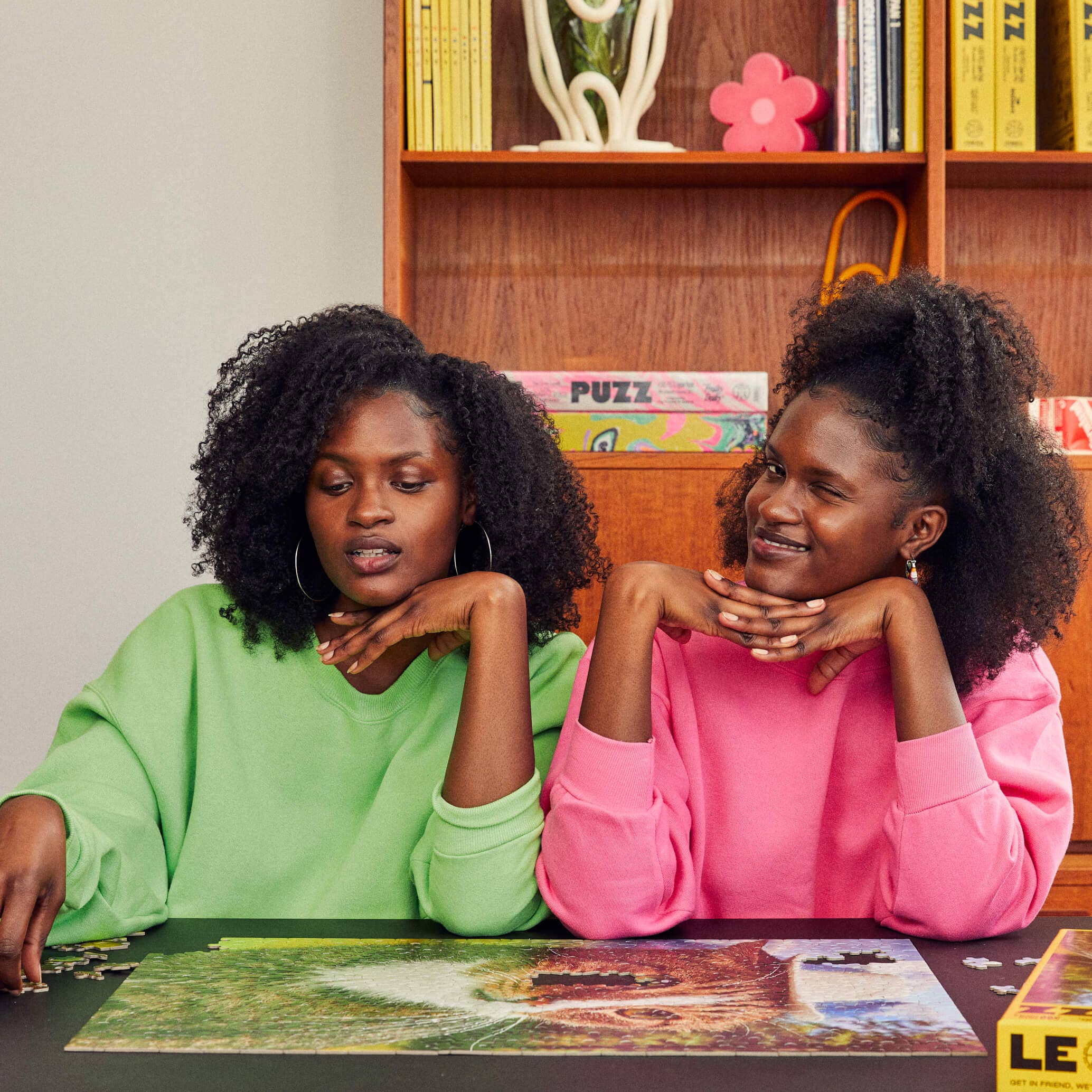 Two women sitting at a table with a puzzle, smiling and looking at the camera.