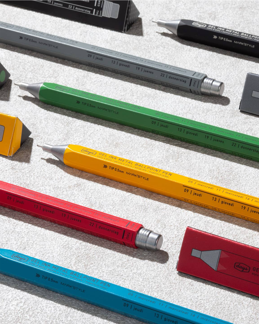 A styled flat-lay of various ballpoint pens in different colors (gray, green, yellow, red, and blue) arranged diagonally on a textured light gray surface.