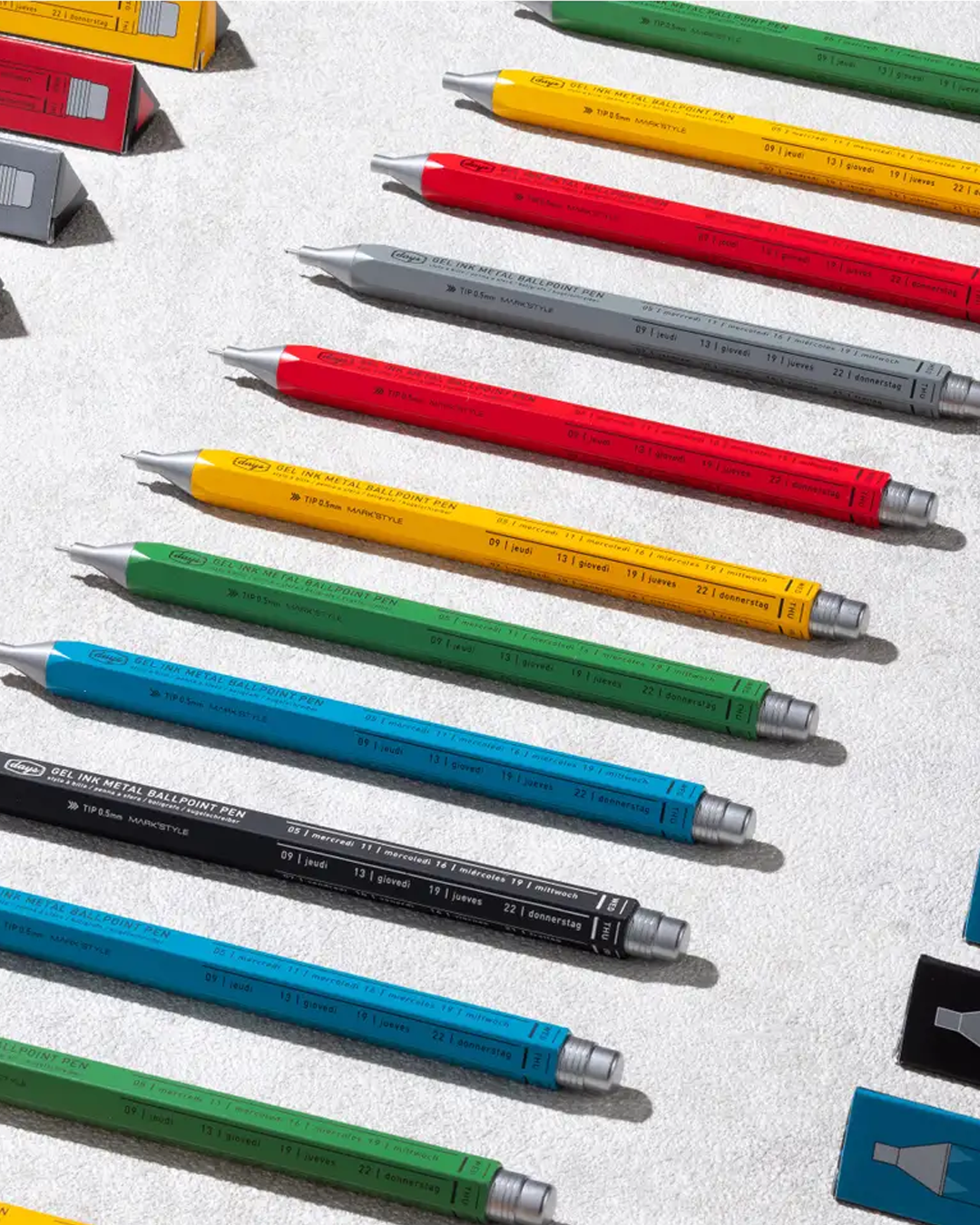 A collection of colorful ballpoint pens in blue, green, yellow, red, and gray, arranged diagonally on a textured light gray surface.