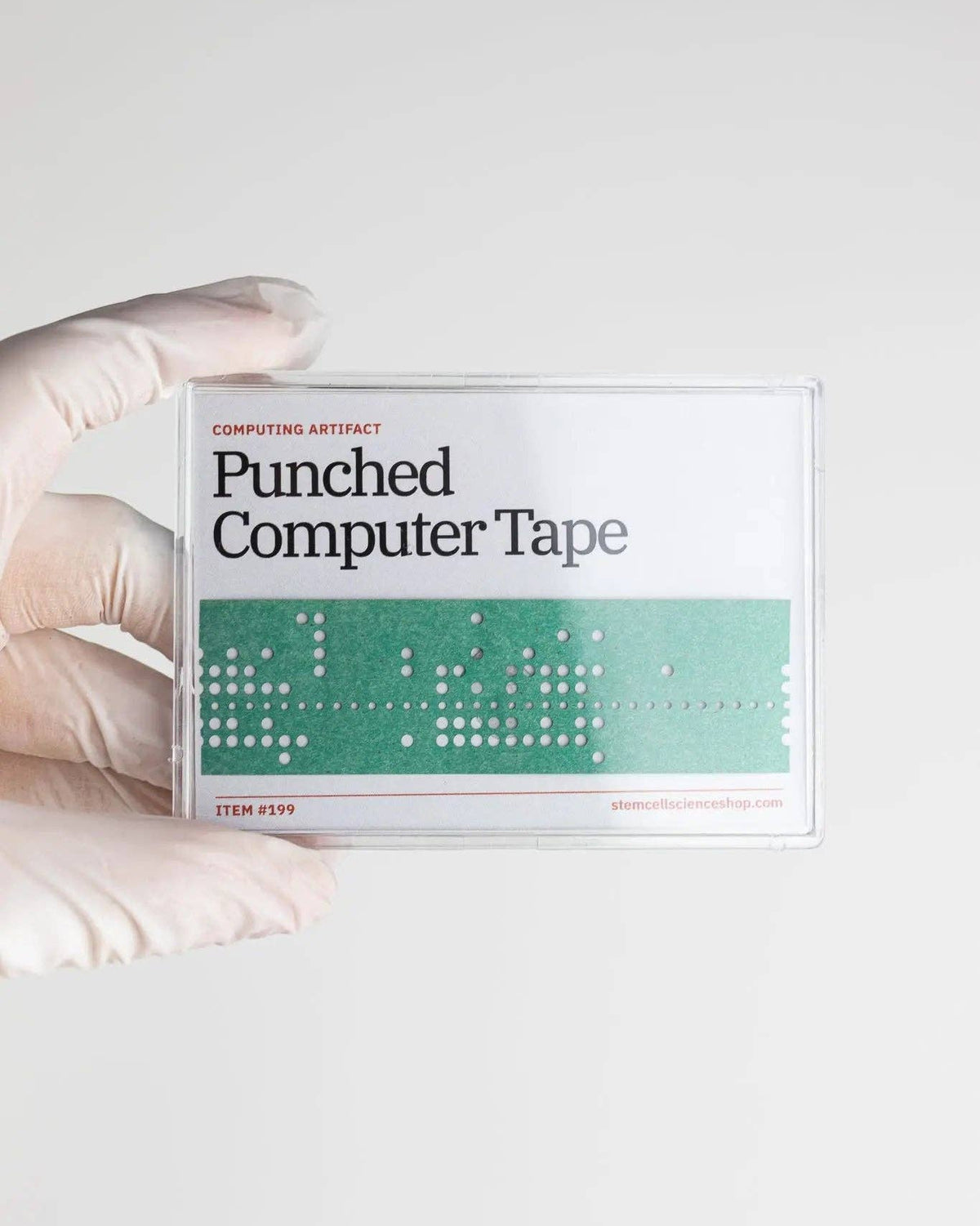 A hand wearing a white glove holds a clear plastic case containing 'Punched Computer Tape' from the Computing Artifact collection against a light gray background.