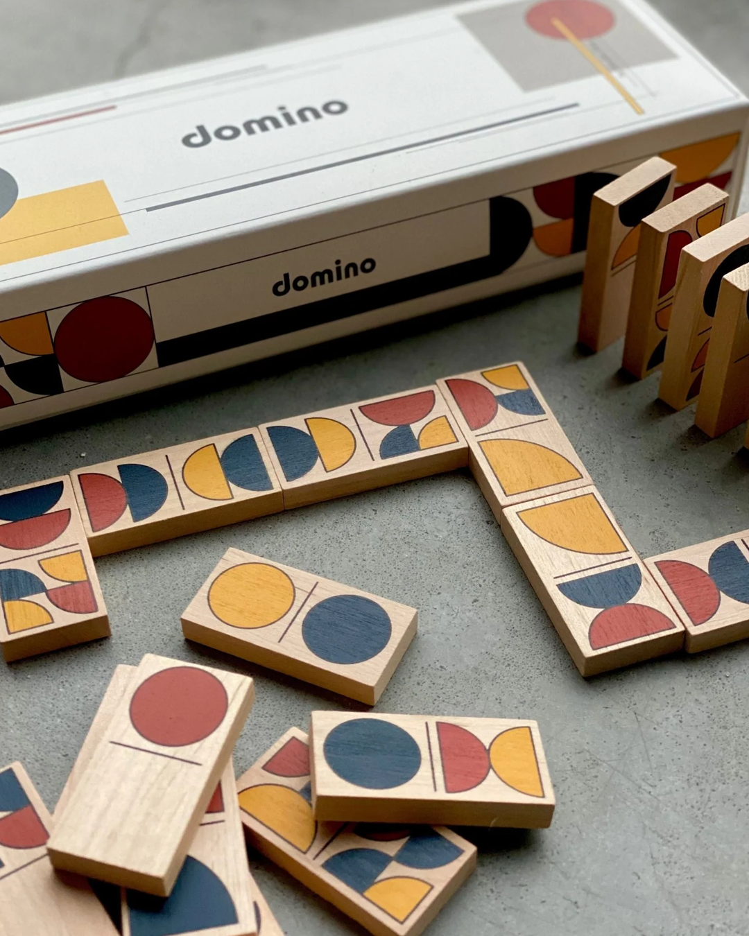 Domino board game pieces with geometric semicircle and circle patterns in yellow, red, and blue, arranged on a gray surface next to their white and black branded box.