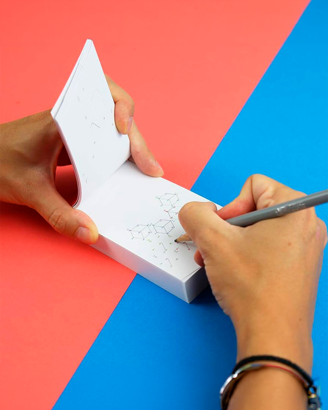 A close-up of a person's hands holding an open white notepad and drawing with a gray pencil, set against a bold diagonally split red and blue background.