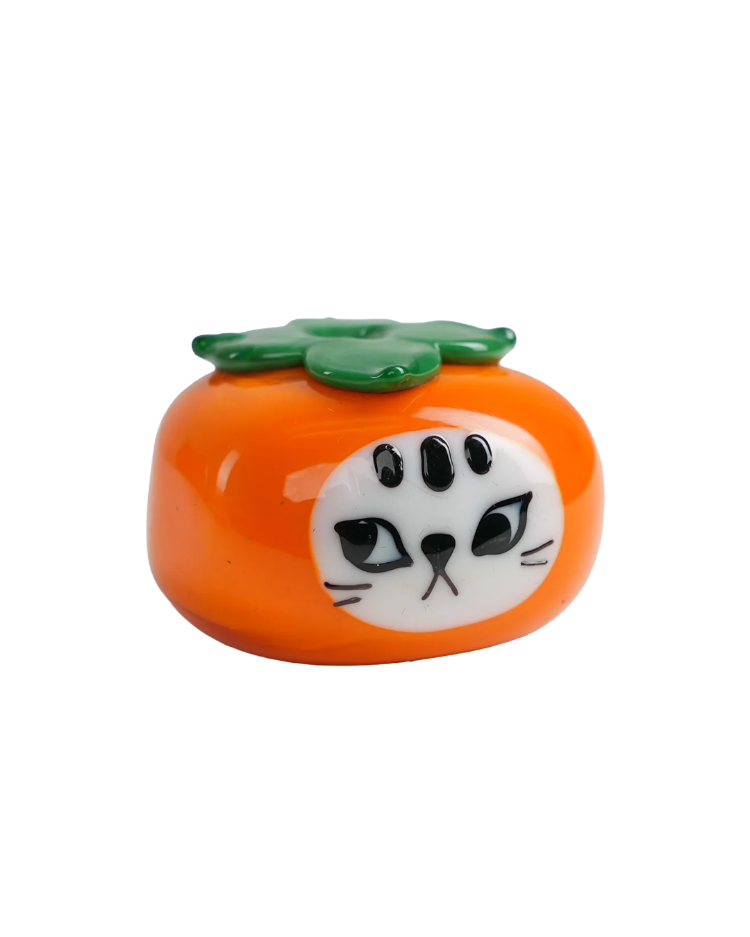 An persimmon-shaped ceramic figurine featuring a white cat face with black eyes, whiskers, and three black spots on its forehead, topped with a green leafy stem, shown on a plain white background.