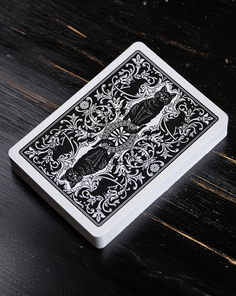 A deck of playing cards with an ornate black and white floral and cat design on the back, resting on a dark wooden surface.