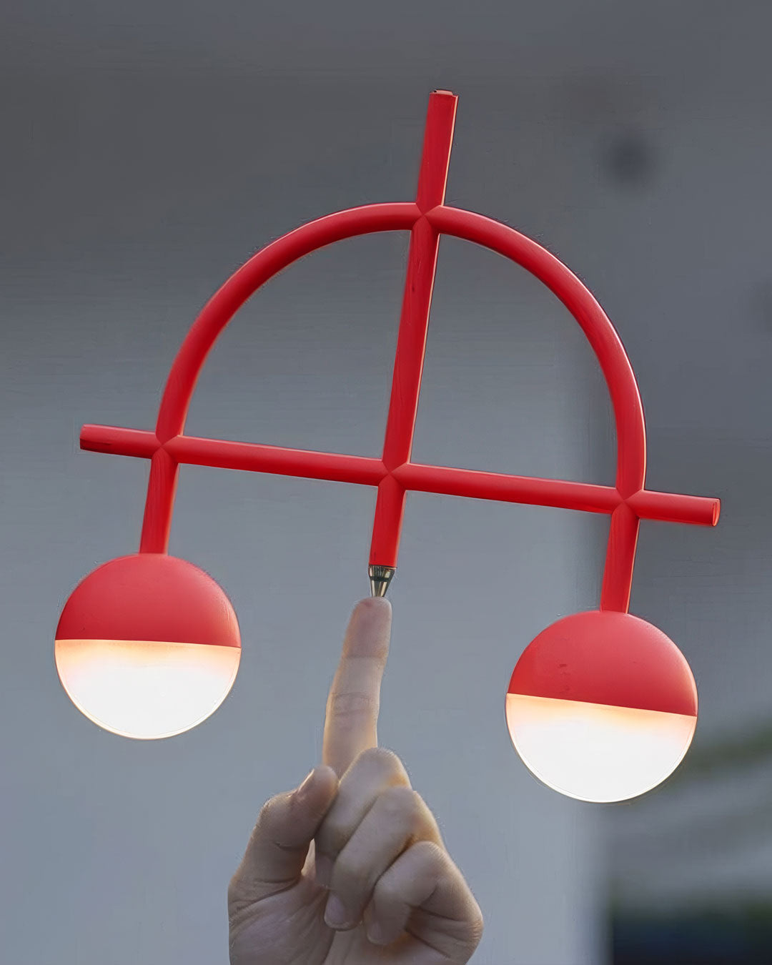 A modern red two-light fixture with spherical glass shades, held by a hand against a blurred gray background.