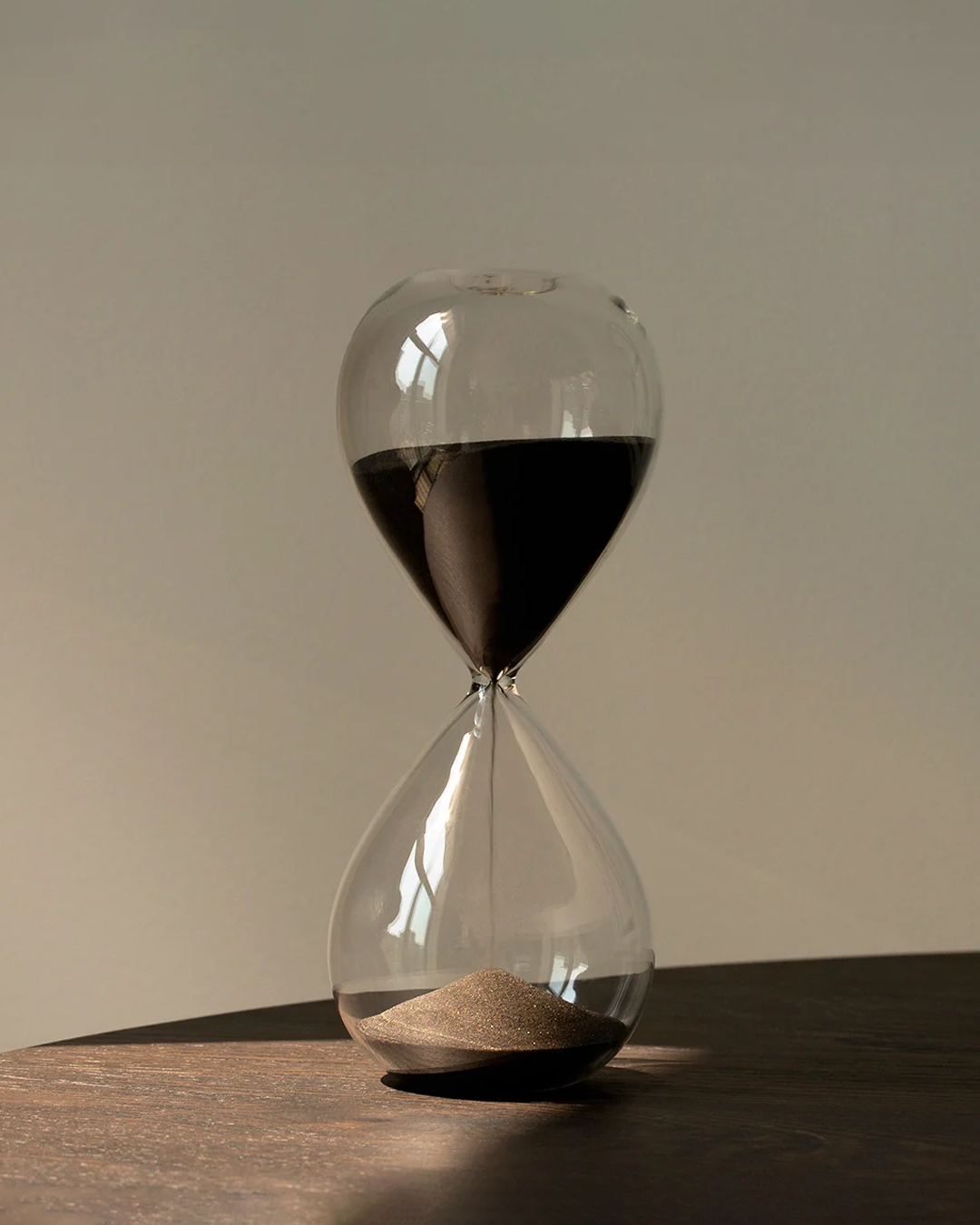 A clear glass hourglass filled with dark sand, sitting on a dark wooden surface against a plain beige background.
