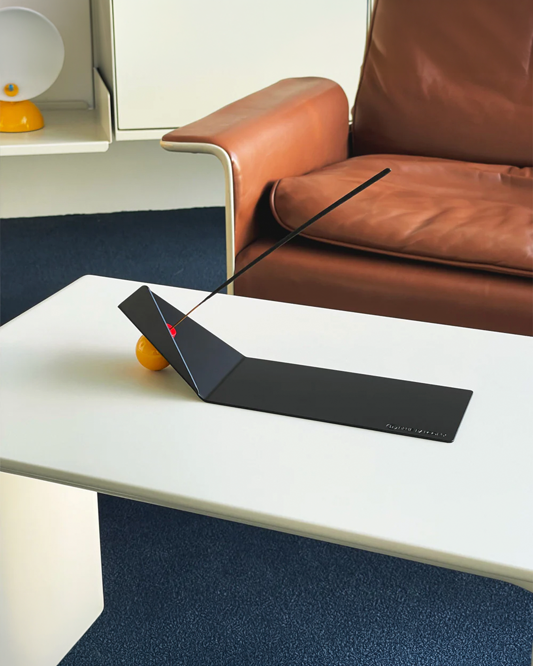 A modern black metal incense holder on a white table in a minimalist room with a brown leather sofa and blue carpet.