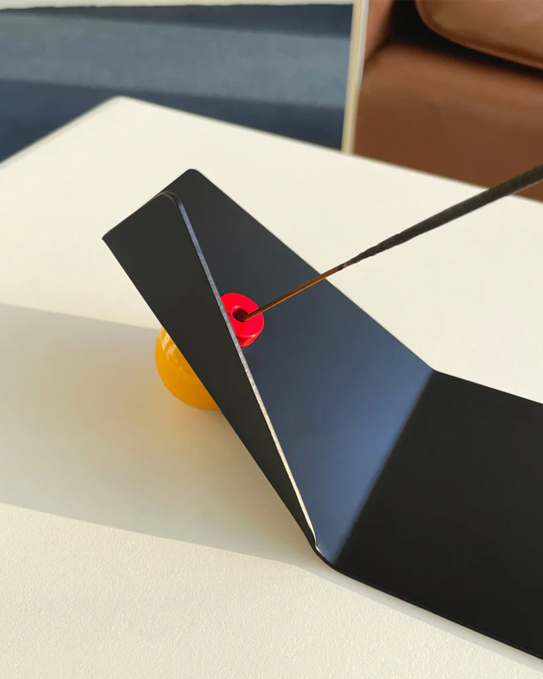 A close-up of a black triangular metal panel on a white surface, with a thin black incense protruding out of a small red circular area near the center. A yellow object is partially visible underneath.