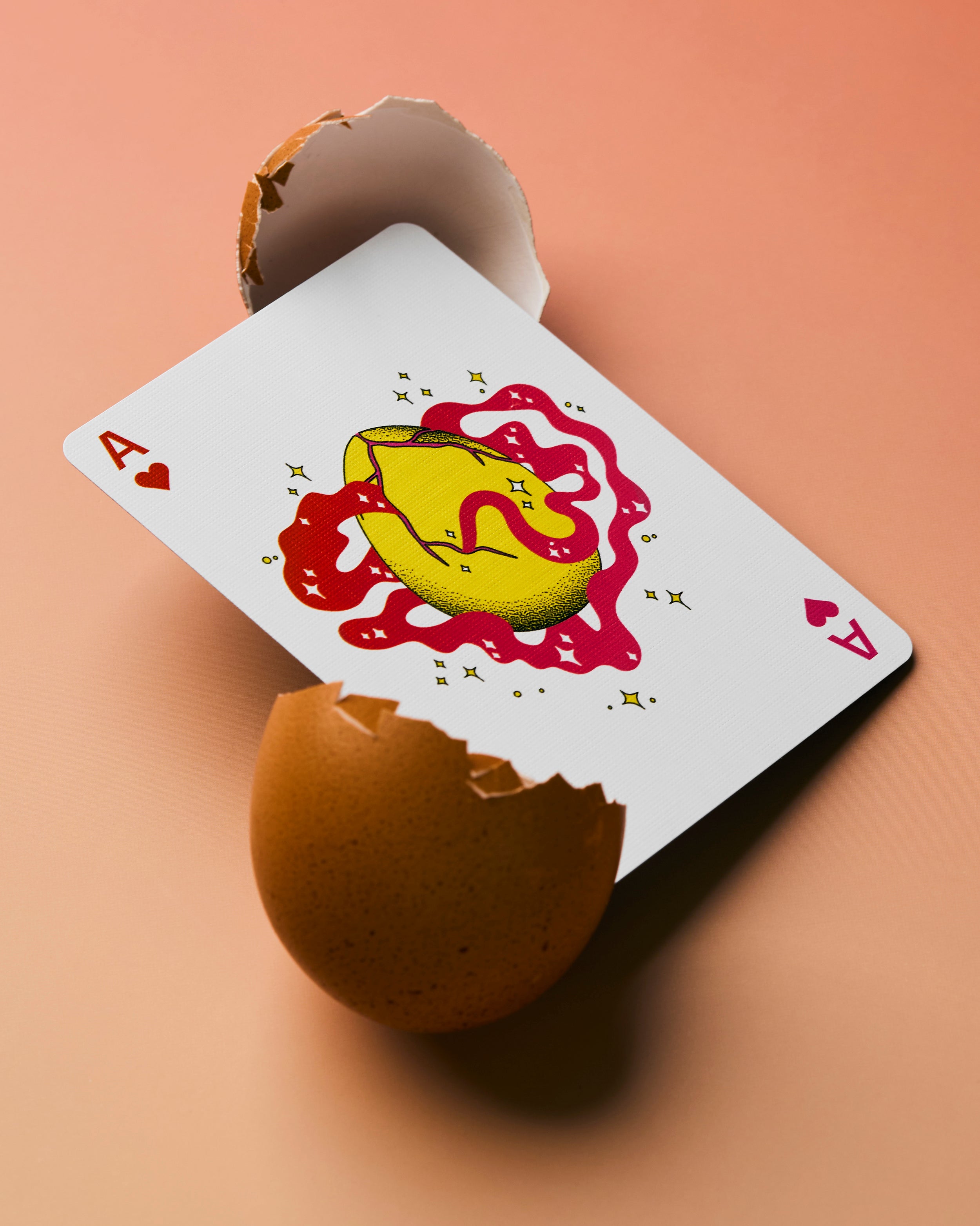 A stylized aces of hearts playing card with a yellow gold ingot and red abstract design, emerging from a cracked brown eggshell on a peach background.