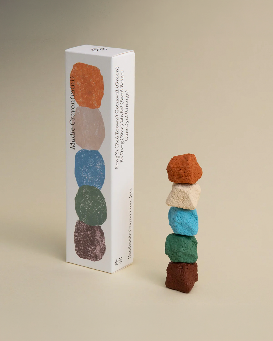 A white product box labeled 'Mudra: Graysonium' stands next to a small stack of six handcrafted crystal stones in orange, cream, blue, green, purple, and brown, photographed on a warm beige studio background.