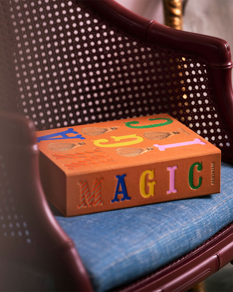 A book titled 'MAGIC' resting on a blue upholstered rattan chair. The book cover is orange-brown with colorful raised letters spelling 'MAGIC' and small illustrated details.